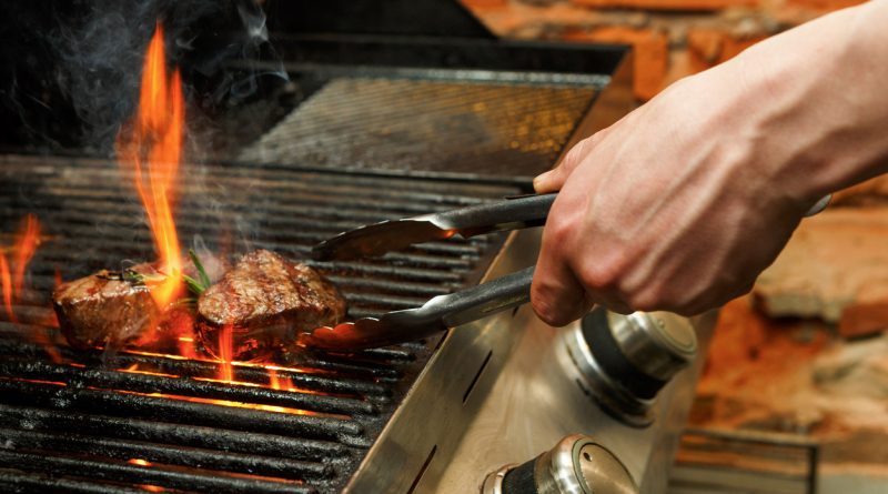 hand using tongs to flip steaks on a grill with flames