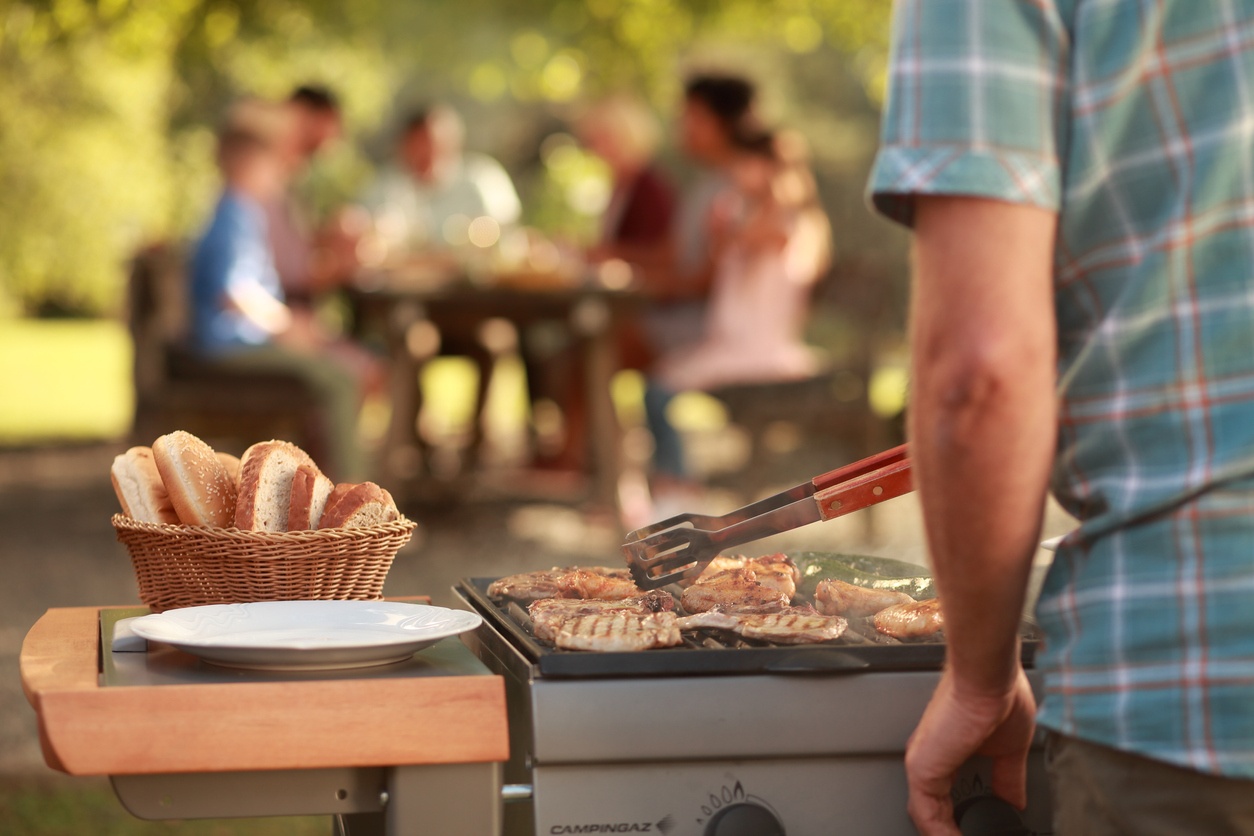 man-in-foreground-right-side-grilling-food-on-gas-grill-with-blurry-people-gathered-in-background-during-party