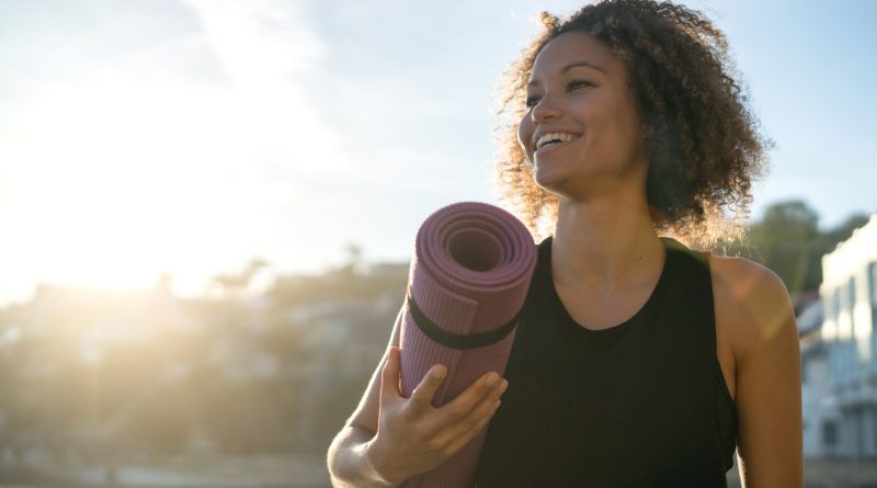 young-woman-outdoors-smiling-while-holding-rolled-up-purple-yoga-mat-under-arm