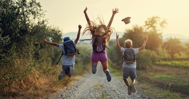 Three kids jumping into the air with their arms up while hiking on a gravel path and wearing camping and hiking backpacks
