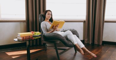 A woman sitting in a reading chair with a high back and low armrests while reading a book with a yellow cover.