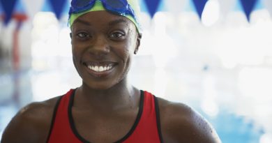 An African American woman wearing a lime green swim cap and a red swim suit smiling at the pool.